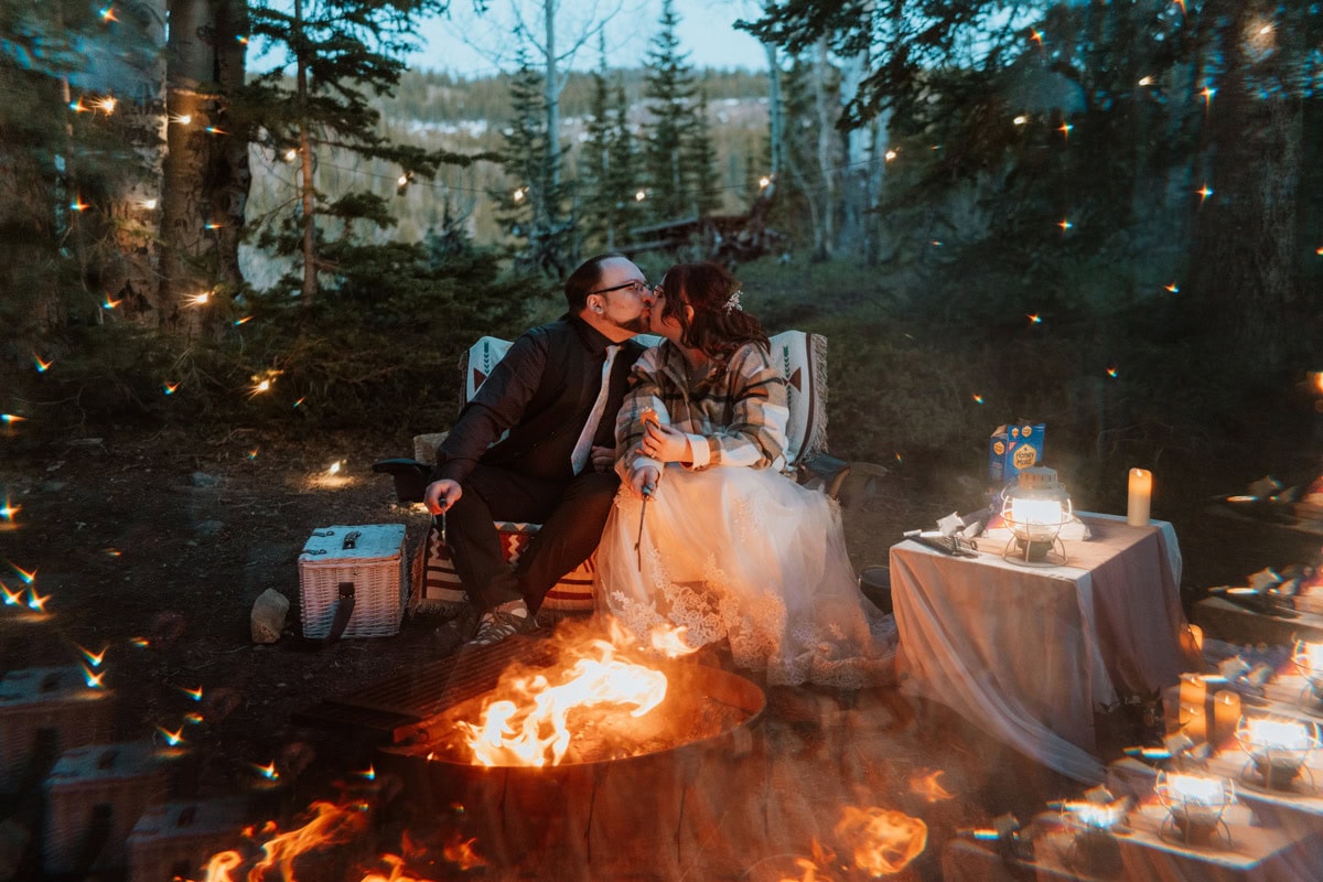 A couple sharing a kiss while sitting by a cozy campfire surrounded by glowing lanterns and string lights during their private mountain destination vow renewal celebration.