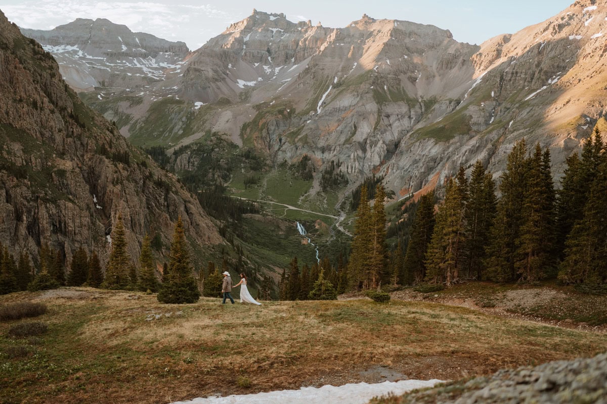 A couple in wedding attire walking through a massive green mountain valley during their Ouray destination vow renewal, with rugged peaks and a distant waterfall in the background.