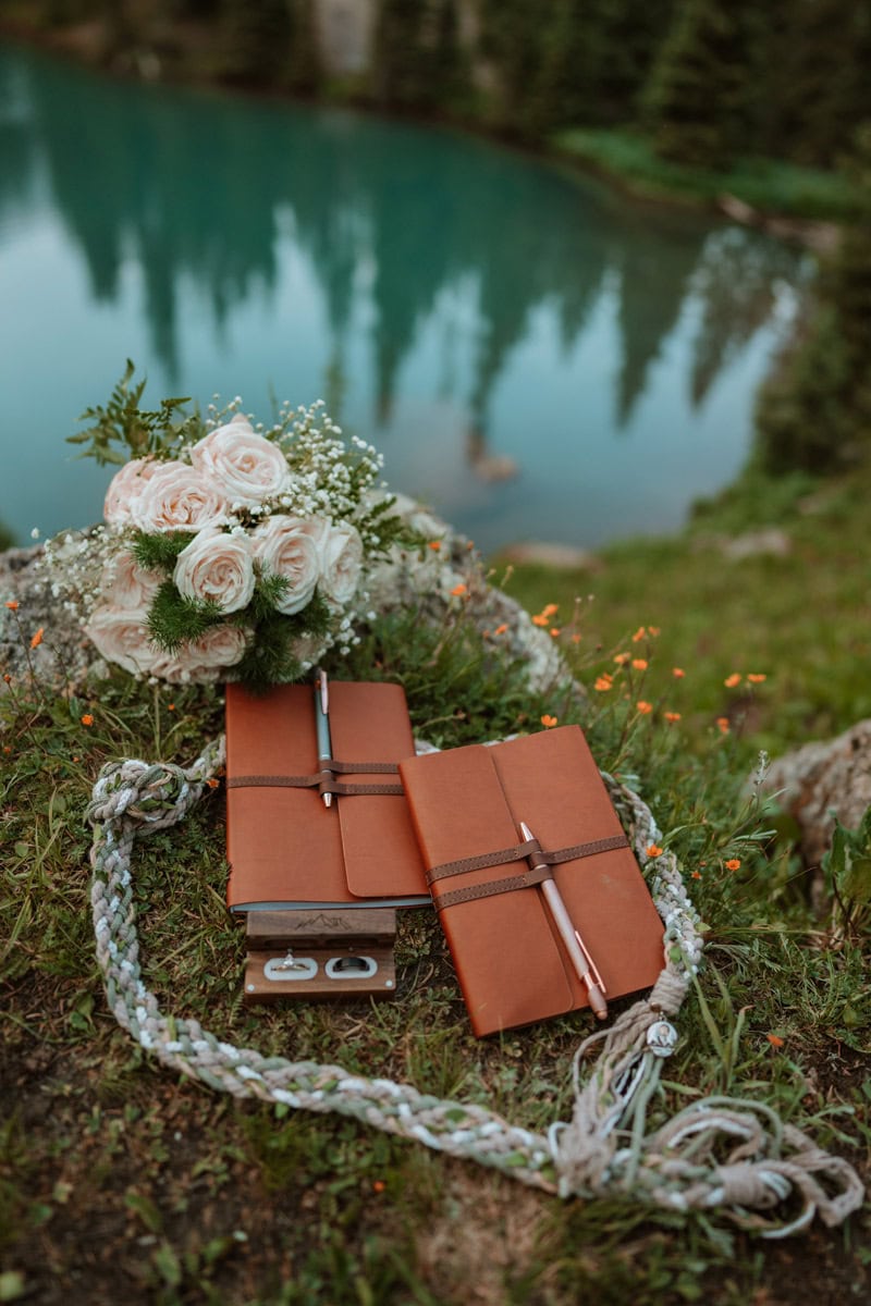 Two brown leather vow books, a white rose bouquet, and a wooden ring box sitting on the grass near an alpine lake for a couple renewing your wedding vows.