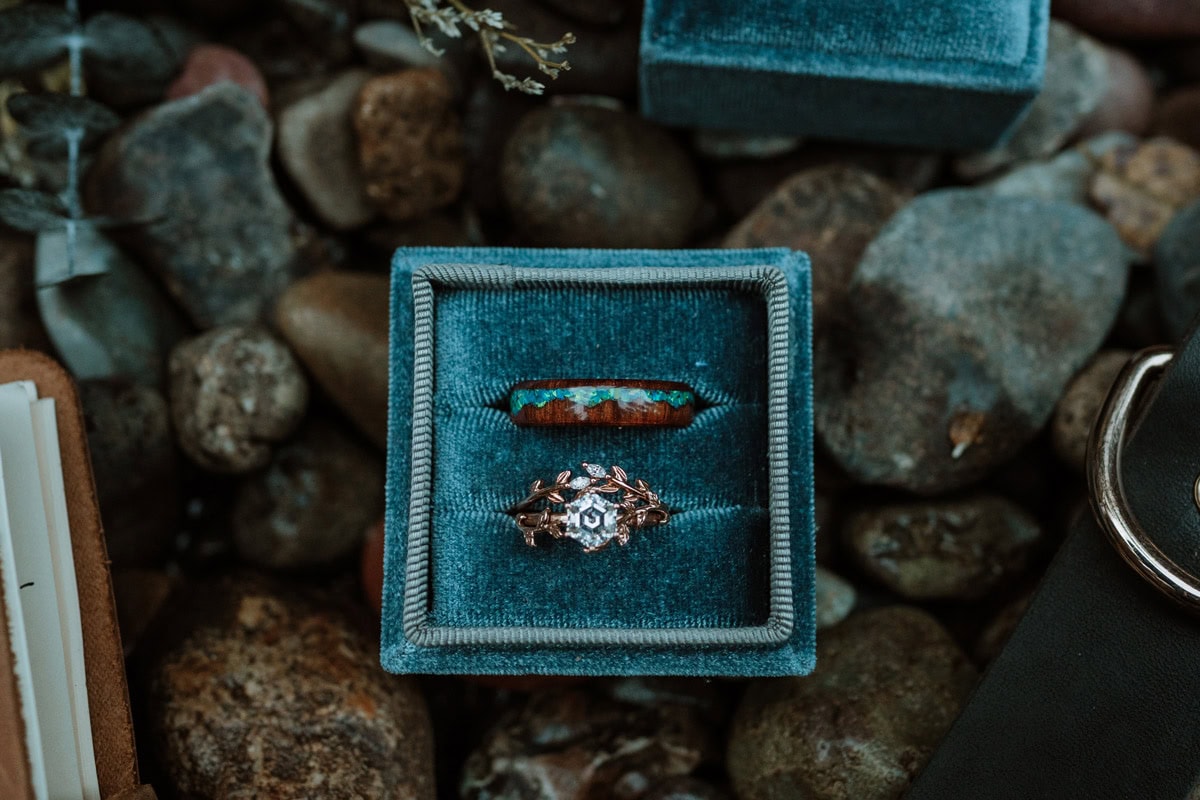 A teal velvet ring box holding a turquoise inlaid band and a delicate diamond ring sitting on river rocks for a marriage renewal vows ceremony.