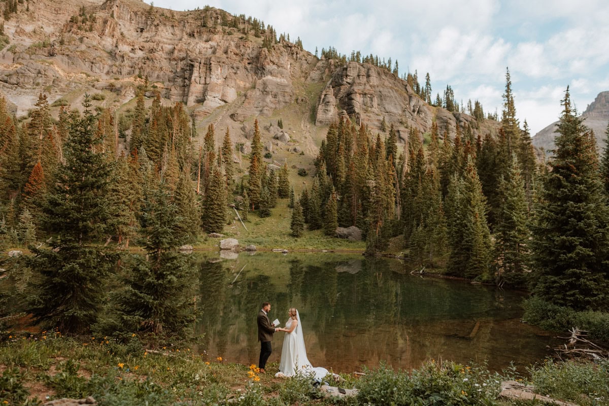 A couple holding hands and reading vows next to a calm alpine lake surrounded by pine trees and rocky cliffs for their summer mountain destination vow renewal in Colorado.