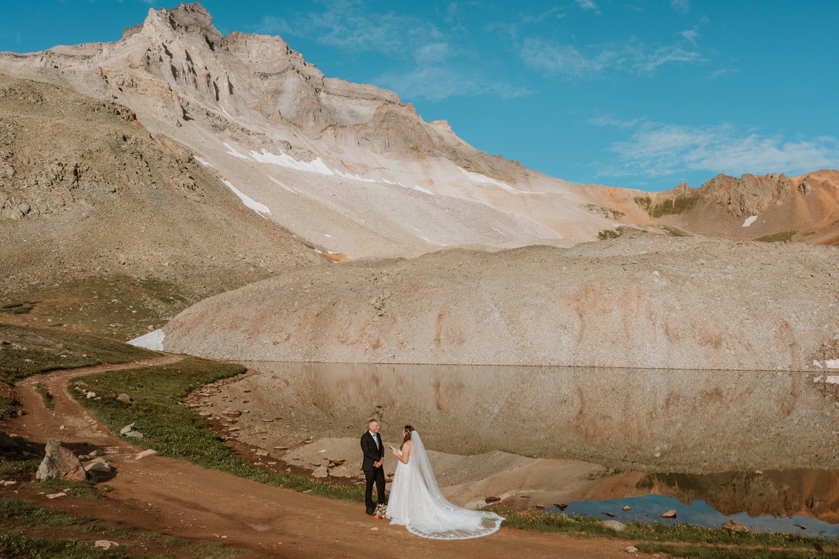 A bride and groom exchanging vow renewal vows at sunrise next to a perfectly still alpine lake reflecting massive rocky mountain peaks.