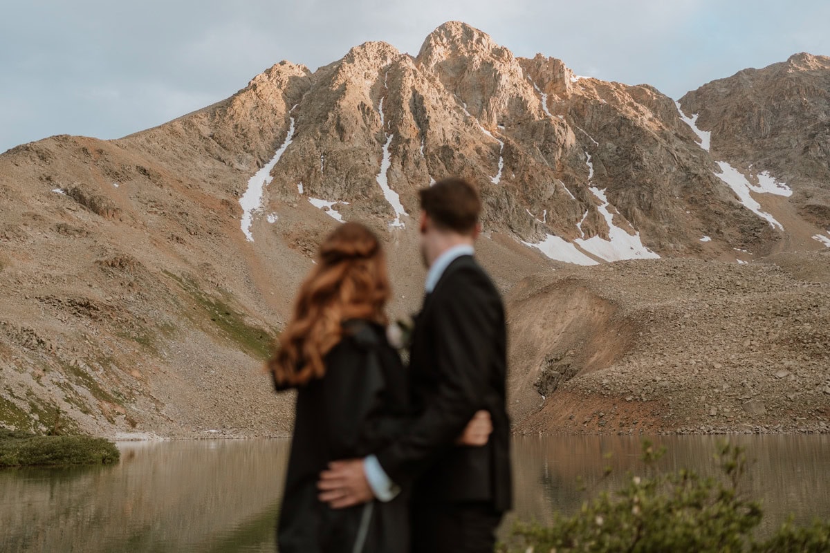 A blurred couple standing by a calm lake, looking up at a massive, sharp mountain peak catching the orange sunset alpenglow during their Aspen destination vow renewal.