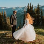 Bride twirls in her wedding dress at sunset. Trees and mountain views tower behind them. The groom is wearing ray ban sunglasses and has a huge smile on his face as he watches his bride twirl.