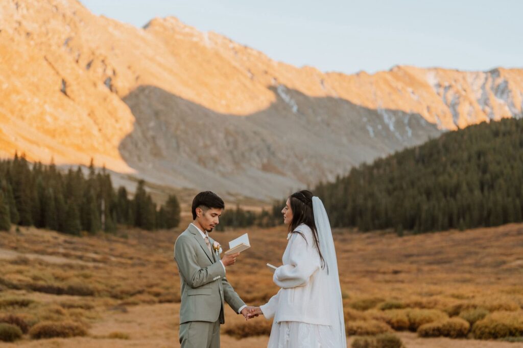 A couple holding hands and reading their vow renewal vows in a golden autumn valley with sunlit mountain peaks behind them in Colorado.