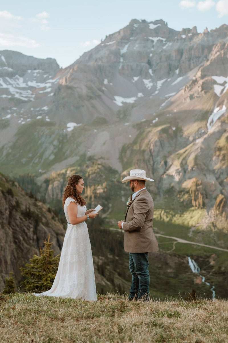 A groom wearing a white cowboy hat and jeans listens to his wife as she reads her renewing marriage vows on a grassy cliff with jagged mountains behind them.