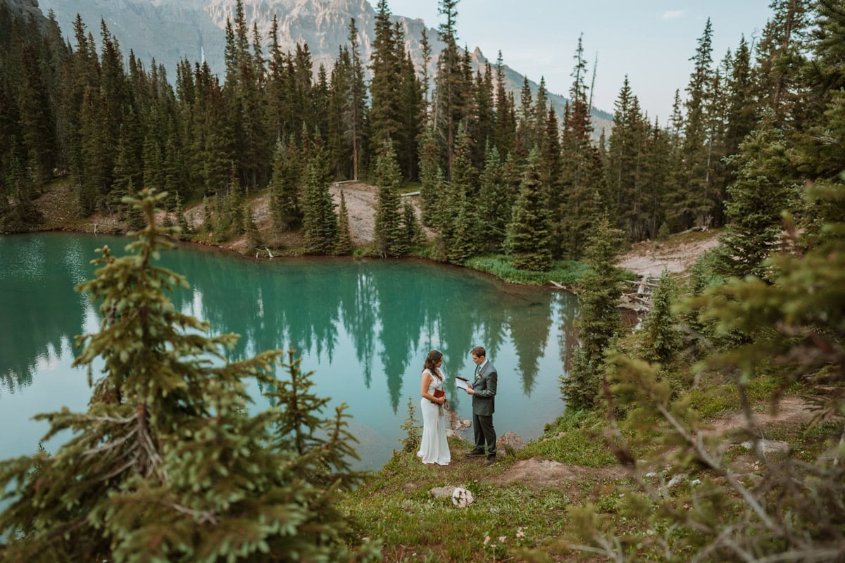 A couple standing on the grassy shore of a vibrant turquoise alpine lake surrounded by evergreen trees while reading their vow renewal vows.