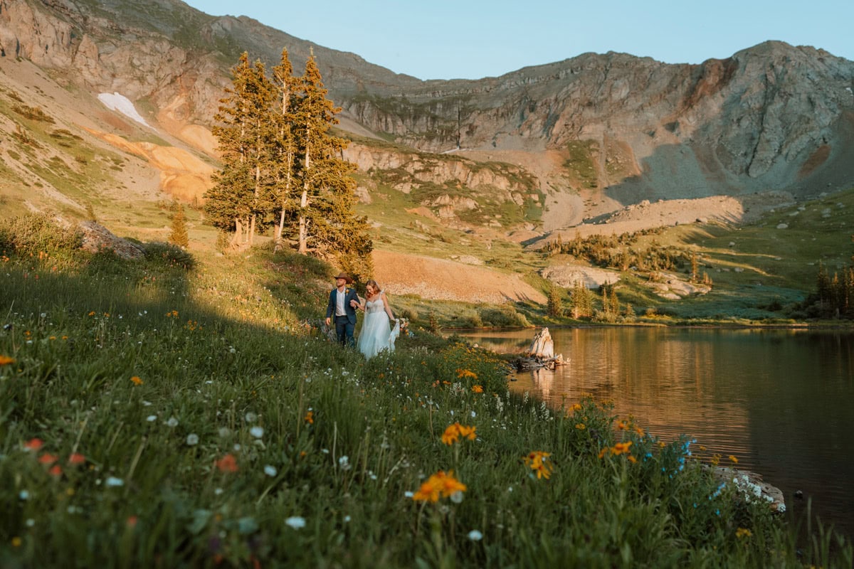 A couple holding hands and walking through a lush green alpine meadow full of colorful wildflowers next to a mountain lake for their Telluride destination vow renewal.
