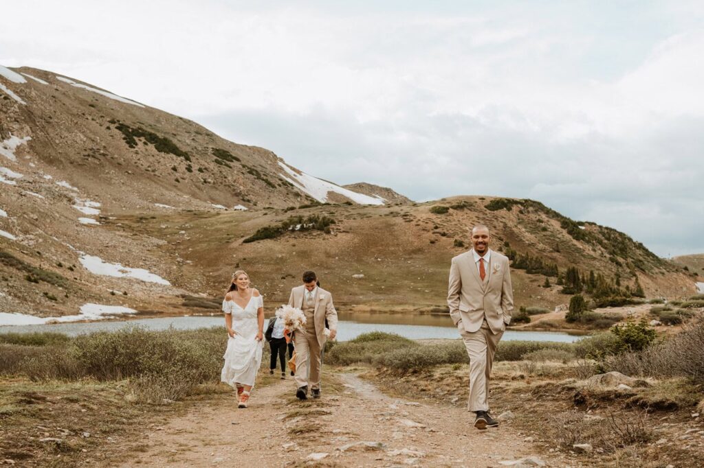 A bride, groom, and a groomsman hike up a dirt trail away from the alpine water after a beautiful elopement ceremony at Loveland Pass under cloudy skies.