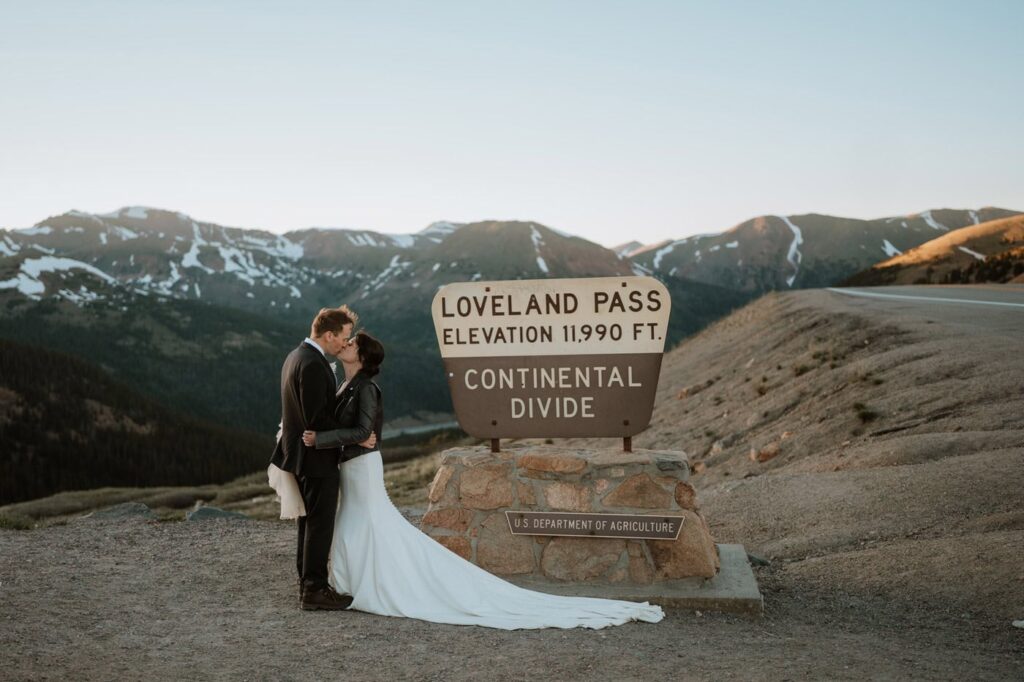A newlywed couple embraces and shares a kiss standing directly in front of the iconic wooden Loveland Pass sign marking the Continental Divide at 11,990 feet for their Loveland Pass elopement.