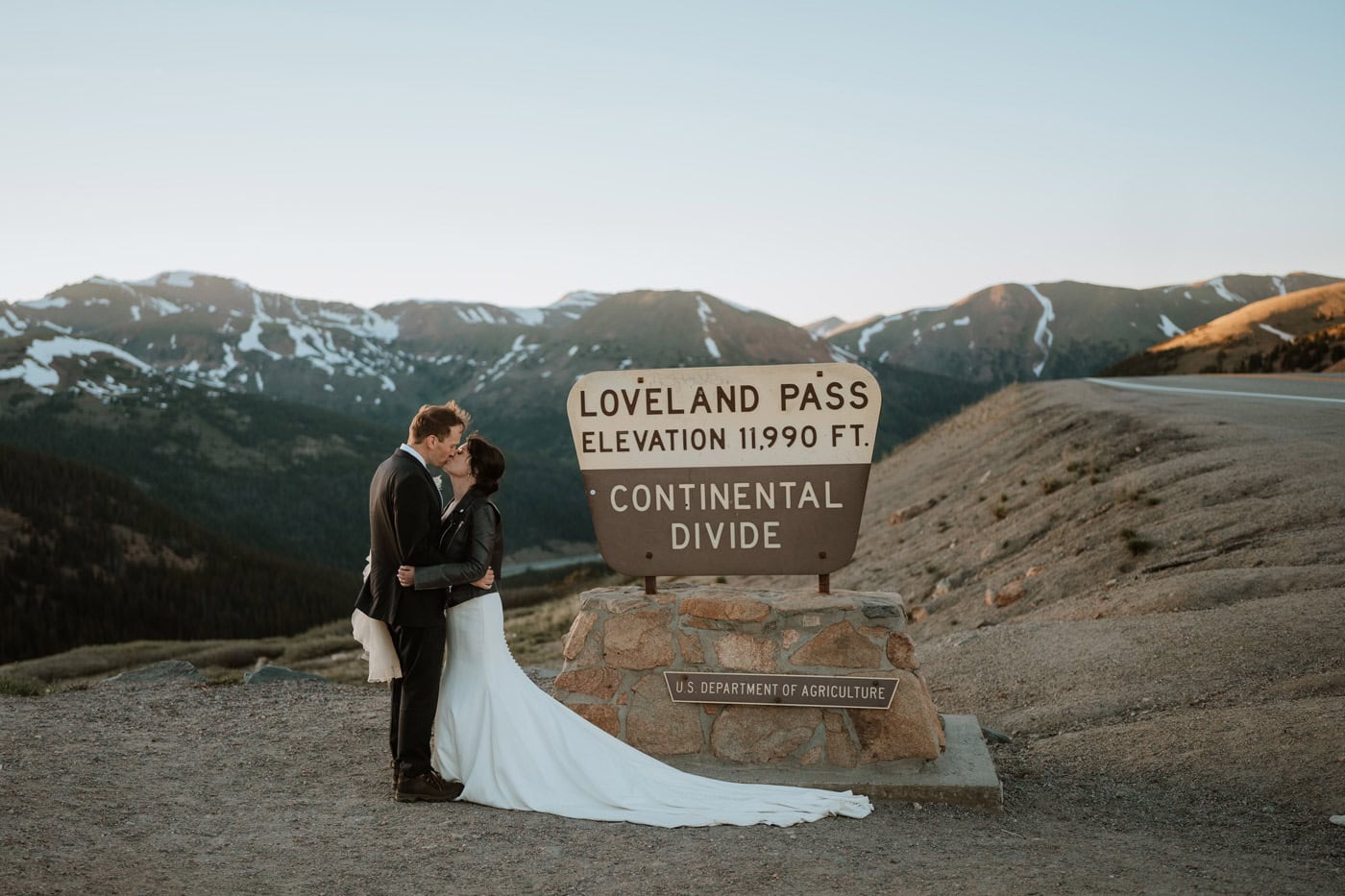 A newlywed couple embraces and shares a kiss standing directly in front of the iconic wooden Loveland Pass sign marking the Continental Divide at 11,990 feet for their Loveland Pass elopement.