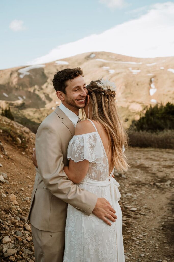 A smiling groom in a tan suit embraces his bride in a beautiful lace dress during their summer Loveland pass elopement, set against a rolling mountain backdrop.