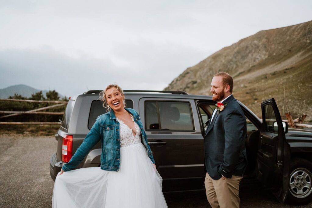A bride wearing a denim jacket over her dress laughs happily beside her groom and their parked SUV, scouting where can you get married at Loveland Pass right off the main road.