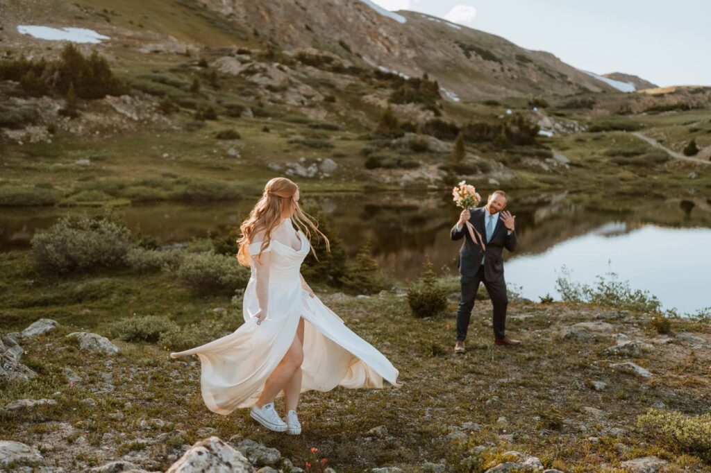 A bride wearing white sneakers twirls her dress playfully as her groom cheers her on holding her bouquet during their Loveland pass wedding by the water.