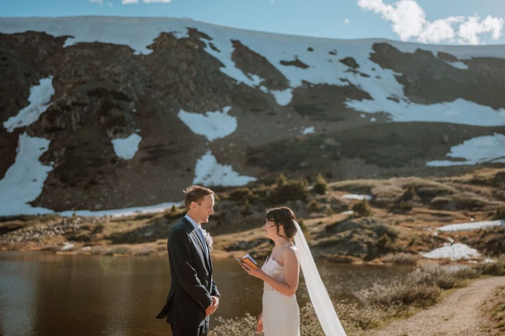 A bride reads her handwritten vows from a small book to her groom during their sunset Loveland pass elopement, backed by a massive mountain slope.