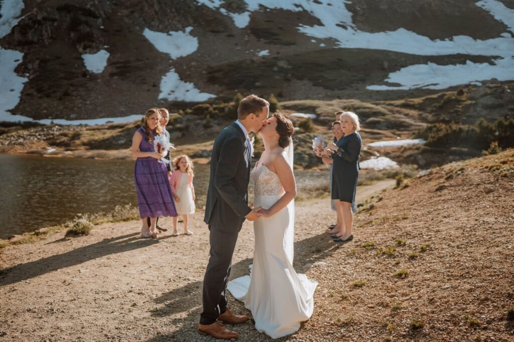 The bride and groom share their first kiss to conclude their lakeside ceremony, celebrating their Loveland Pass Lake wedding while their family smiles in the background.