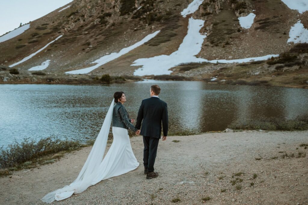 A bride wearing a leather jacket over her dress walks hand-in-hand with her groom toward a scenic Loveland pass lake beneath snowy mountain peaks.