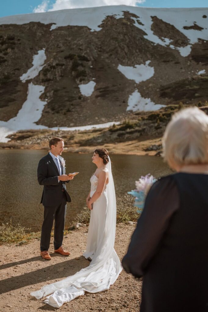A groom reads his vows from a small leather booklet to his smiling bride by the water while getting married at Loveland Pass.