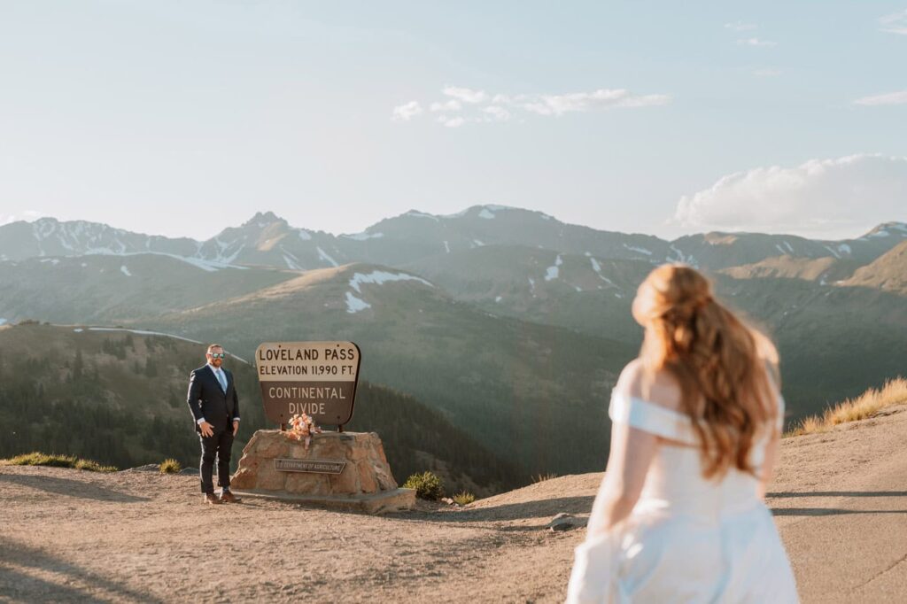 A groom smiles at his approaching bride while standing next to the iconic Loveland pass sign marking the Continental Divide at 11,990 feet.