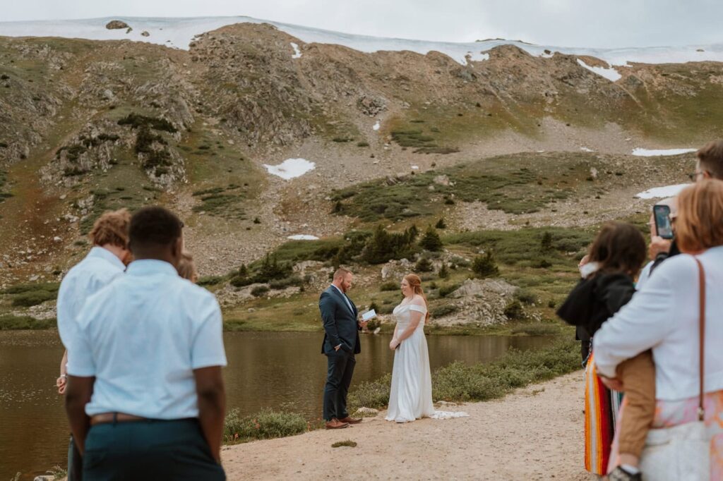 A groom in a navy suit reads his vows to his bride while standing near an alpine lake with their guests during a beautiful wedding ceremony at Loveland Pass.