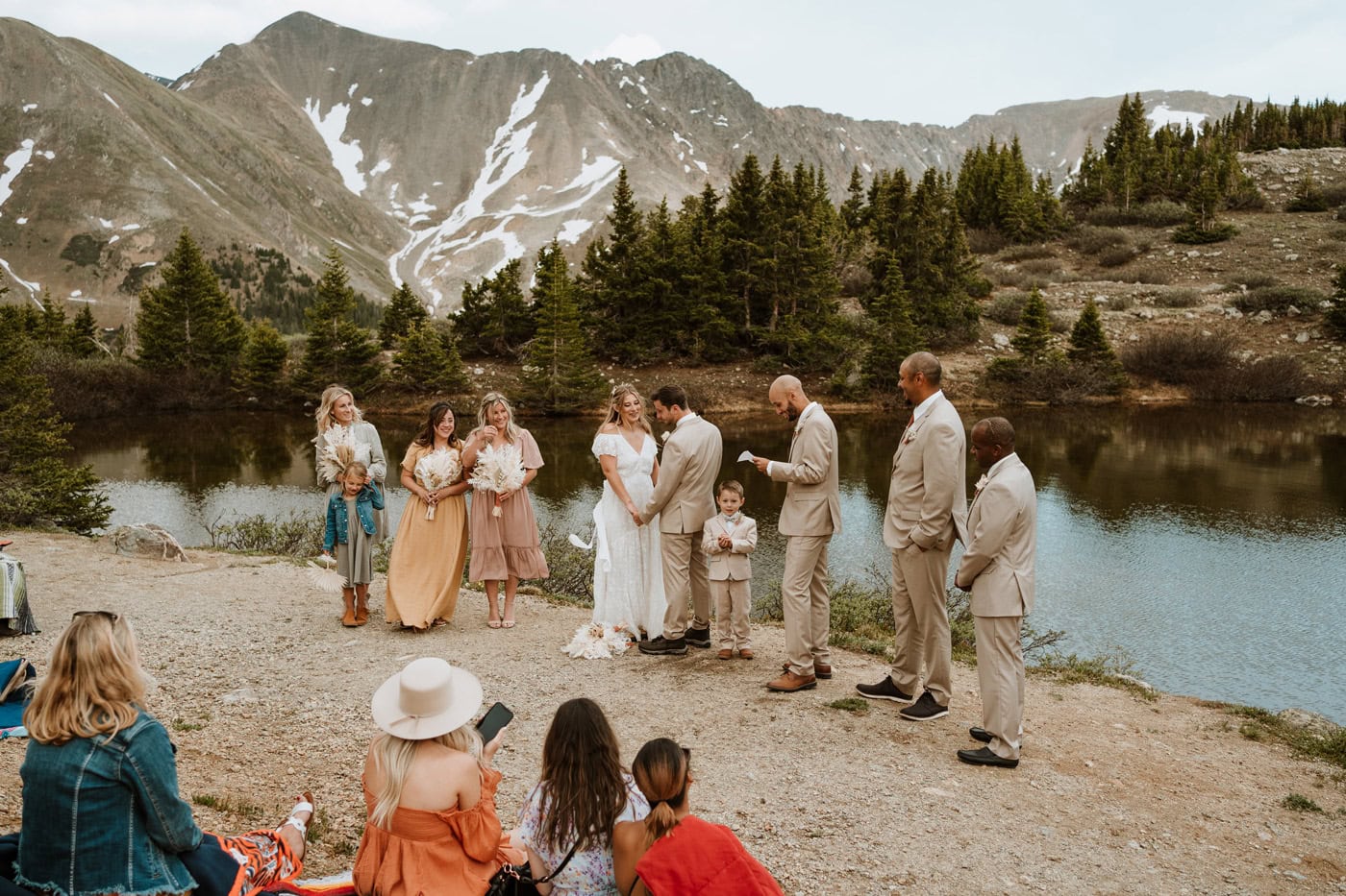 A bride and groom hold hands during a Loveland Pass wedding ceremony, surrounded by family gathered near the alpine water.
