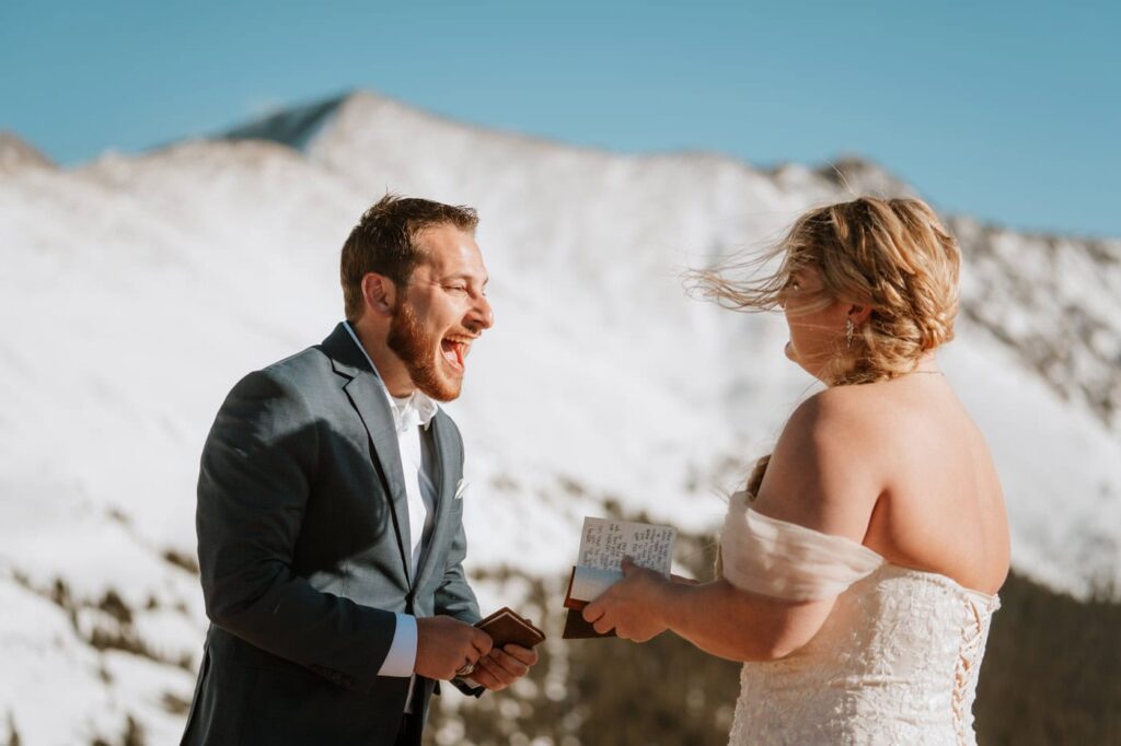 A groom laughs joyfully as his bride reads from leather vow books in the wind while getting married at Loveland Pass surrounded by bright white snow.