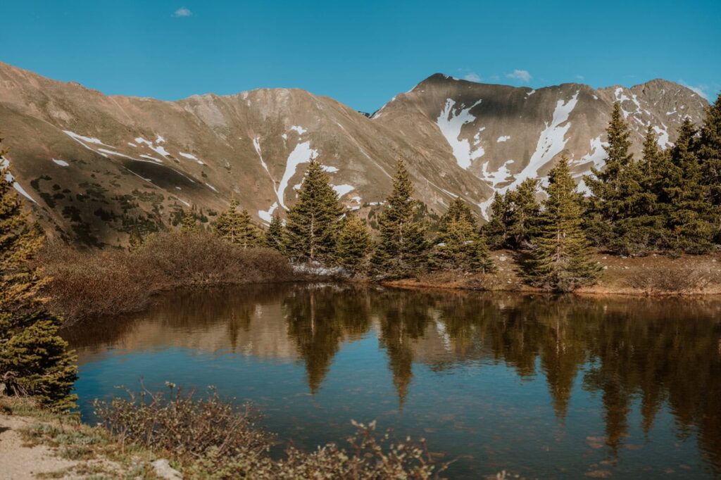 Evergreen trees and steep, snow-dusted mountains reflect perfectly in the calm, clear waters of Loveland Pass Lake on a sunny afternoon.