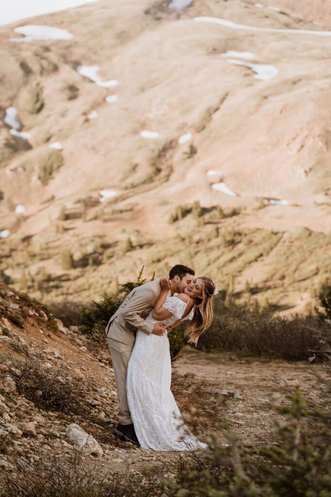 A groom leans in to kiss his laughing bride's cheek on a mountain slope, celebrating their scenic Loveland Pass Colorado wedding.