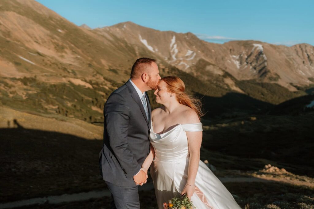 A groom sweetly kisses his bride's forehead bathed in golden hour light at the top of Loveland pass, with sweeping mountain peaks behind them.