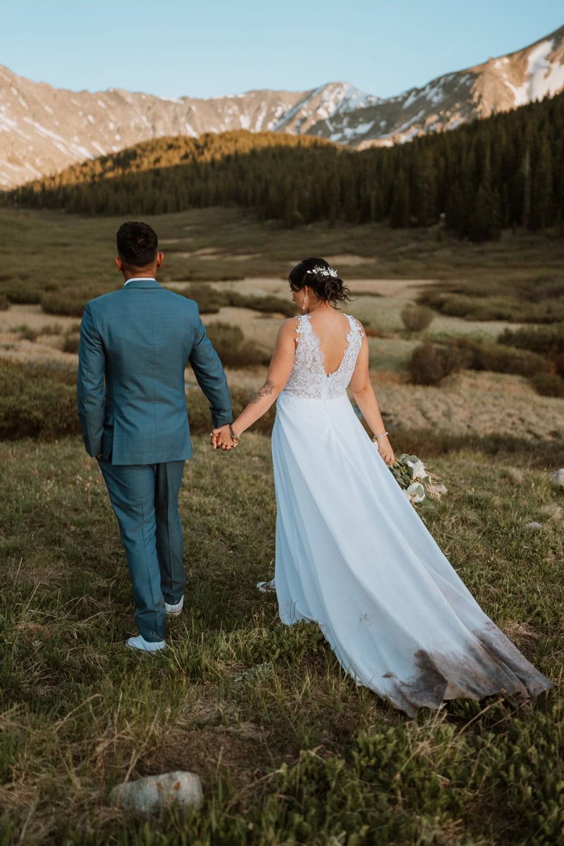 A bride and groom hold hands and walk through an alpine valley, highlighting a badge of honor dirt on wedding dress elopement idea.